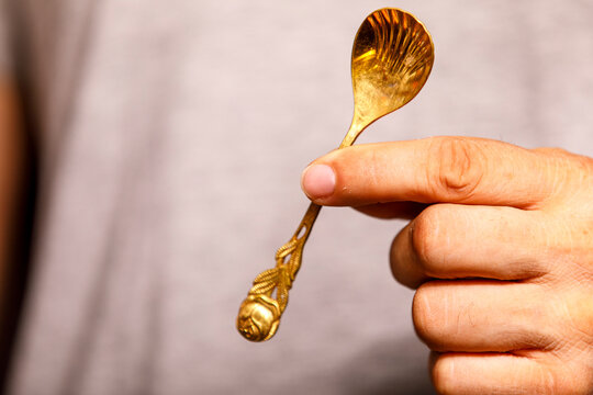 A person's hand holds a small, intricately designed golden spoon against a light background.
