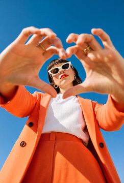 Woman in orange outfit forms heart shape with hands against clear blue sky