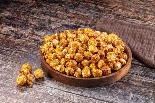 Caramel popcorn in wooden bowl on rustic wood surface with scattered kernels and brown cloth napkin