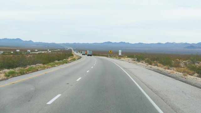 Pov shot driving on long straight desert modern highway in american prairie with sparse traffic and mountains visible on horizon under cloudy sky during daytime. Interstate transportation goods