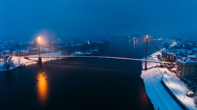 Aerial view of the illuminated St. Irenaeus Bridge spanning the Sava River at dusk with snow-covered banks in Sremska Mitrovica, Vojvodina, Serbia.