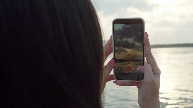 Close-up of a woman recording a video of the sunset over the sea using a smartphone.