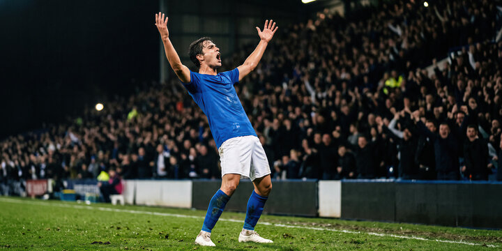 Victorious soccer player raises both arms before packed stadium crowd with overwhelming joy. Winning moment, athletic achievement concept.