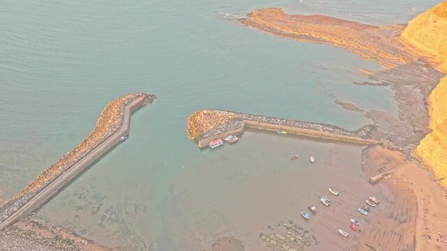Staithes harbour shows twin protective breakwaters, fishing boats, tidal mudflats and rocky shoreline beneath Cowbar Nab on the North Yorkshire coast, drone forward flyover