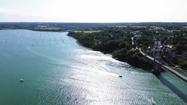 rone shot over the Rance estuary, revealing Pont Saint-Hubert and a peaceful Breton village in the distance. Dozens of sailboats float across the shimmering blue-green water under a clear summer sky.