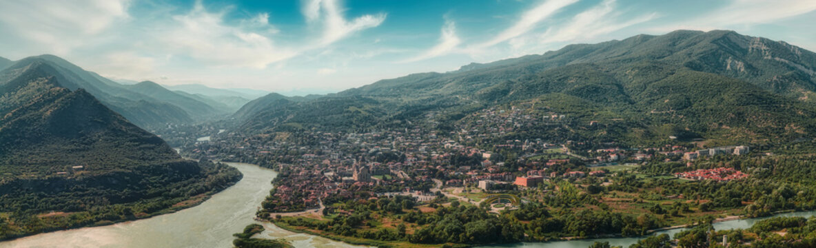 Wide aerial panorama of historic Mtskheta town at the confluence of rivers surrounded by Caucasus mountains under blue sky with clouds in Georgia.