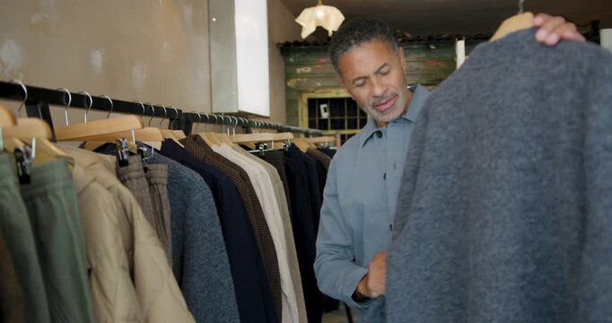 Mature Man Shopping for Clothes in Store, Exploring a Collection of Cozy Sweaters