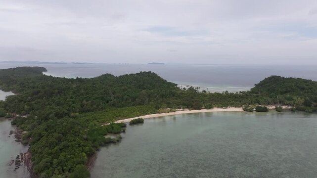 Aerial view of Batam Island featuring lush green hills, a pristine sandy beach, and clear shallow waters under a cloudy sky in Batam, Riau Islands, Indonesia.
