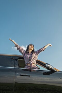 A joyful woman leans out of a car window, stretching her arms wide in celebration. She is surrounded by a clear blue sky and lush fields, embodying the spirit of adventure on a sunny day