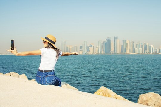 Excited Woman traveler visitor sit raised arms at waterfront enjoy Doha skyline viewpoint across bay symbolizing freedom urban lifestyle travel Middle East modern city concept