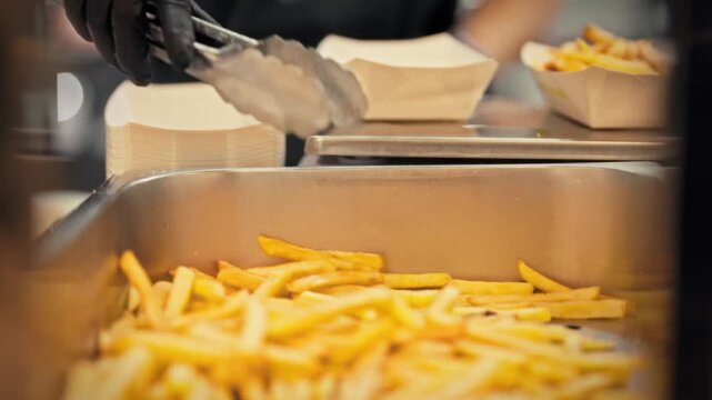 A cook in an apron and gloves places cooked French fries into a container in a bright fast food kitchen. Concept for professional preparation and serving of fast food takeout dishes