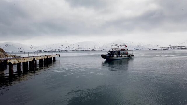 Akdamar Island pier on Lake Van, Turkey, with a tour boat arriving in winter and snowcapped mountains behind, the ferry to the Armenian Church of the Holy Cross