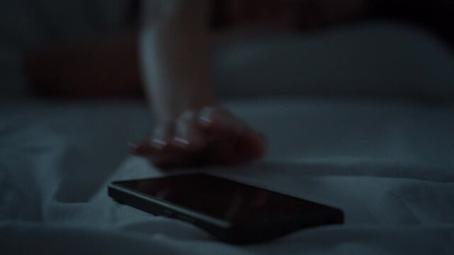 A smartphone lies near the hand of a sleeping girl who reaches out automatically during the night, capturing the pervasive theme of modern technology and mobile device dependence idea