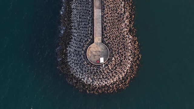 Aerial drone view of a circular breakwater pier extending into calm blue ocean waters, showing geometric tetrapod rock armor, textured coastline and a solitary walkway bathed in warm evening light