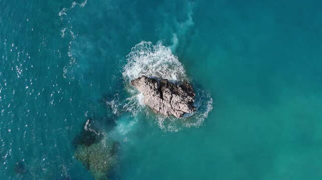 Aerial view of a jagged rock surrounded by turquoise sea water and white foam waves in Cyprus.