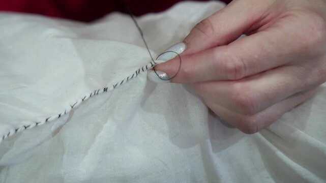 Hands sew white fabric by hand with a needle and black thread in close-up