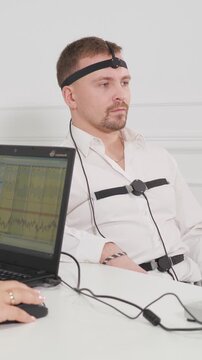 Nervous man connected to polygraph sensors answering questions during a lie detector examination, with a specialist monitoring his vital signs on a laptop computer in a white room