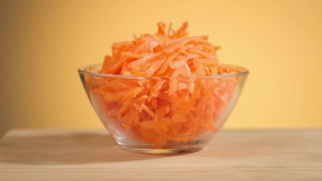 Freshly grated carrots in salad glass bowl against orange background, closeup of vegetables, preparing healthy dieting vegan meal