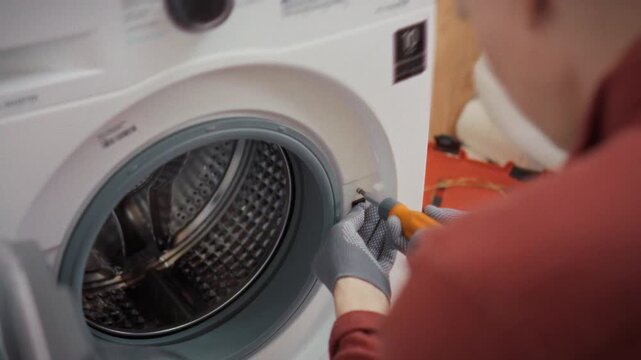 An experienced technician in work uniform unscrews bolts from a washing machine body in a bathroom with a toolbox in the background. Concept of professional repair and replacement of appliance parts