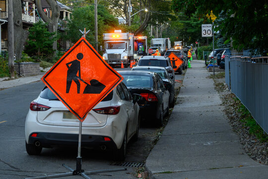 Road work sign and utility trucks with traffic control flaggers on a tree-lined residential street at dawn in Toronto, Ontario, Canada