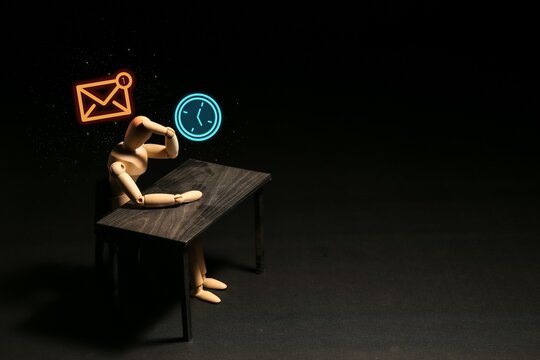 Stressed wooden mannequin sitting at a desk under glowing email and clock icons, capturing a real photo of deadline anxiety, overwork, and digital burnout.