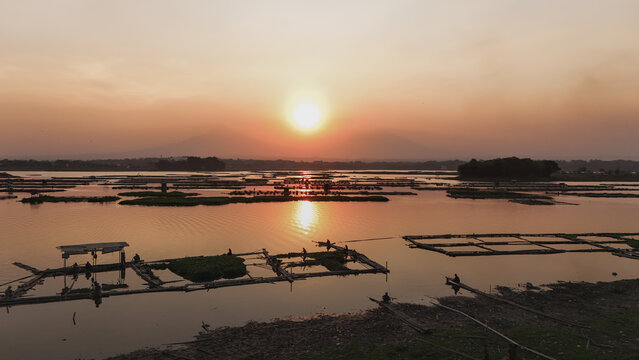 Aerial view of Cengklik Reservoir at sunset with floating fish farms and fishermen, featuring Mount Merapi and Mount Merbabu in the background in Ngemplak, Central Java, Indonesia.