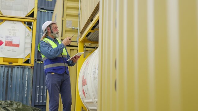 An experienced port worker in a hard hat uses a digital tablet for inventory management and freight inspection at an industrial shipping container yard.