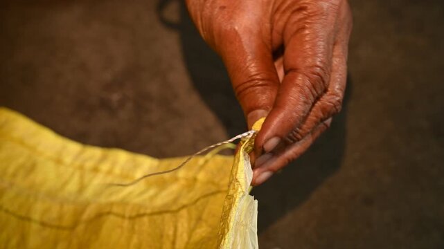 An elderly woman sews a rice sack by hand with thread and needle. Heavy-duty needles and thick plastic thread are commonly used tools for sealing bulk transport sacks. Wholesale markets (mandis)