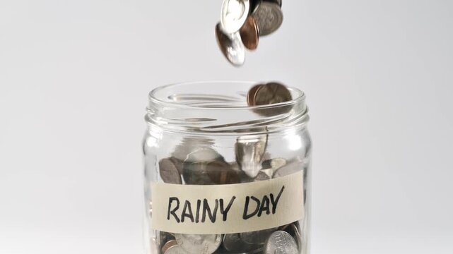 Close-up shot of coins falling in slow motion into a coin jar labeled 'Rainy Day'.