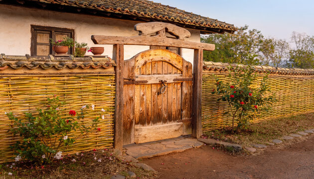 Rustic wooden gate with bamboo fence and flowering bushes outside house.