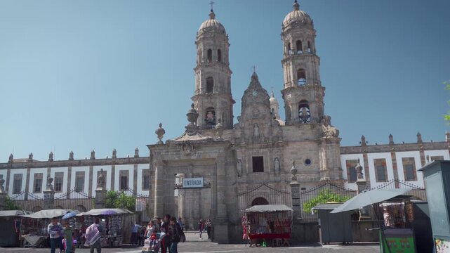 Basilica of Our Lady of Zapopan facade featuring colonial architecture, twin towers, central entrance, surrounding white walls with windows, and visitors walking under clear blue sky