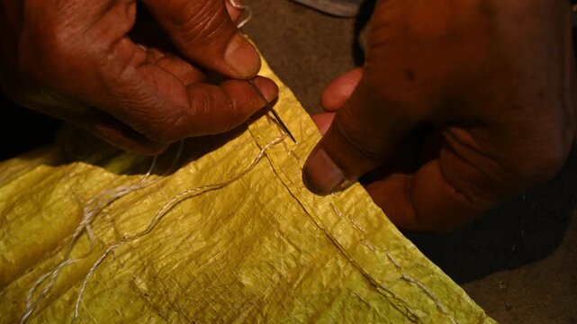 An elderly woman sews a rice sack by hand with thread and needle. Heavy-duty needles and thick plastic thread are commonly used tools for sealing bulk transport sacks. Wholesale markets (mandis)