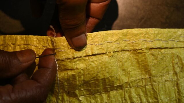 An elderly woman sews a rice sack by hand with thread and needle. Heavy-duty needles and thick plastic thread are commonly used tools for sealing bulk transport sacks. Wholesale markets (mandis)