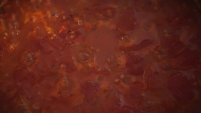 Close-up overhead shot of homemade tomato sauce simmering in a metal pot. Chunks of tomato pulp and bubbles visible on the surface. Idea of traditional sauce cooking at home
