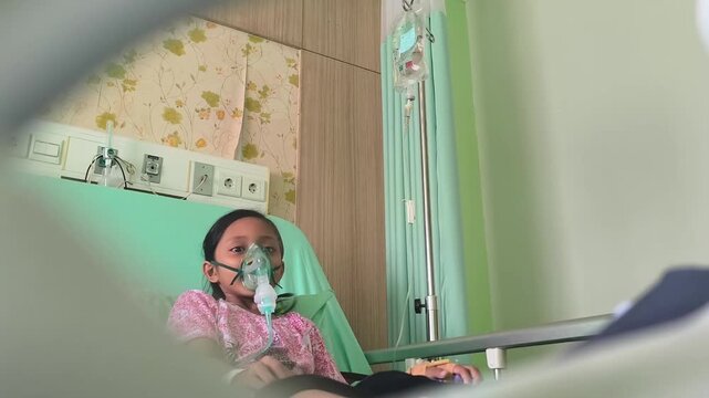 A little girl is using a steam inhalation nebulizer on a hospital bed, looking relaxed