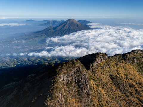 Aerial view of Mount Sumbing rising above a sea of clouds with rugged volcanic terrain and lush green valleys in Kecamatan Kalikajar, Jawa Tengah, Indonesia.