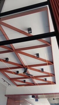 Interior ceiling features exposed reddish-brown wooden beams forming geometric panels, recessed black spotlights, linear lights, and a central dark ceiling fan.