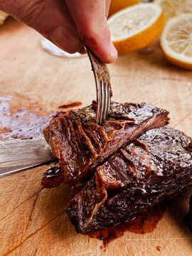 Hand using a fork to separate tender, juicy beef short ribs, cooked on an Asado grill, resting on a wooden cutting board with sliced lemons, representing a traditional gourmet meal