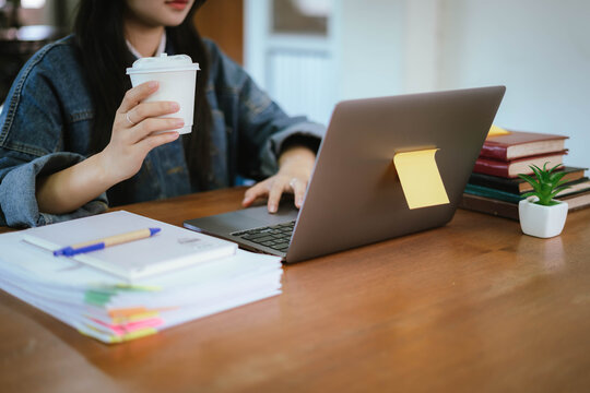 Smiling young woman studying at a desk with a laptop, notebook, pen and takeaway coffee in a bright modern workspace. Concept of online learning, education, productivity, remote study, and student lif