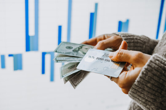 Hands holding US dollars and a bank card in front of the stock exchange screen showing trading graphics.