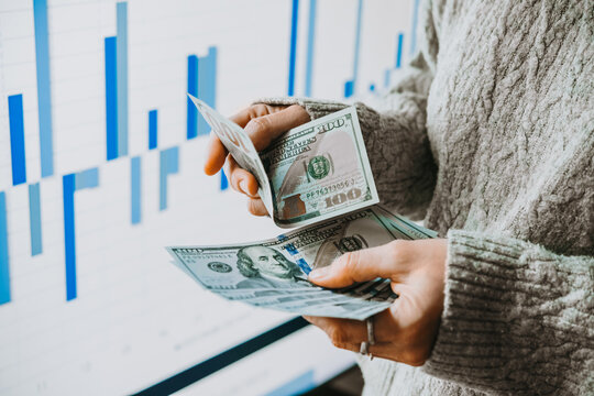 Hands holding US dollars currency in front of the stock exchange screen showing trading graphics.