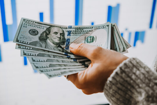 Hands holding US dollars in front of the stock exchange screen showing trading graphics.