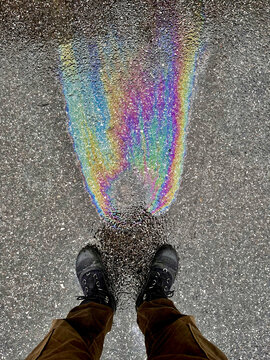 A high-angle point-of-view photograph looking down at a person's feet, standing on asphalt next to a patch of iridescent oil residue reflecting a rainbow spectrum of colors.