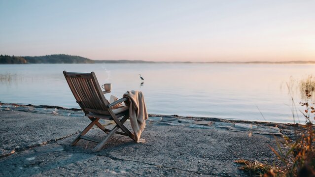 Wooden deck chair with blanket on a rocky shore beside a calm misty lake at golden hour