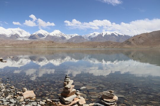 Kala Kule Lake, China 07-09-2026. Stone Cairns on the Rocky Shore of Kala Kule Lake with Perfect Reflection of Snow-Capped Muztagh Ata Mountain Peaks.
