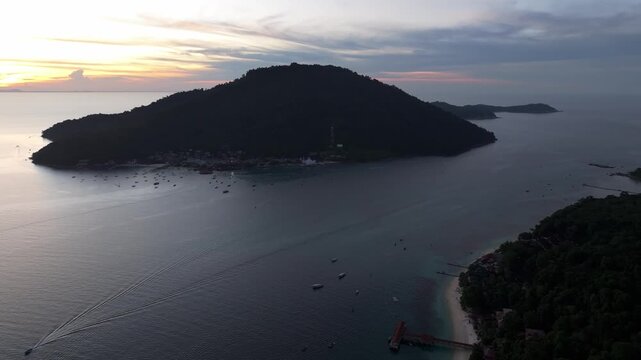 Aerial view of Perhentian Islands at sunset with a boat leaving a wake in the calm sea, lush tropical vegetation, and a wooden jetty in Terengganu, Malaysia.
