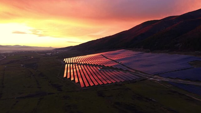 Red sunset light bathes a hillside solar farm, Bulgaria. Warm tones color one panel section while shaded rows and a valley village recede into dusk.