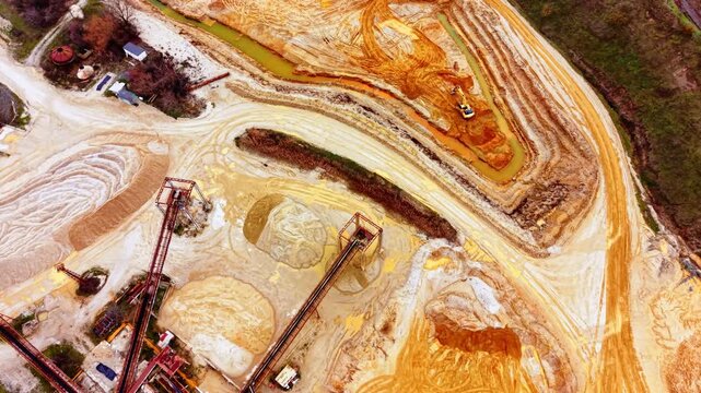 Excavator works beside an orange sediment channel. Aerial view captures swirling mineral stained water beside conveyor towers and sand piles.