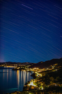 Aerial view of the illuminated coastal town of Corfu at night with star trails across the sky and city lights reflecting on the calm Ionian Sea, Corfu, Ionian Islands, Greece.