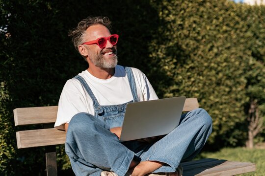 Happy mature man in denim overalls and red sunglasses working on a laptop on a park bench for outdoor remote work and lifestyle banner